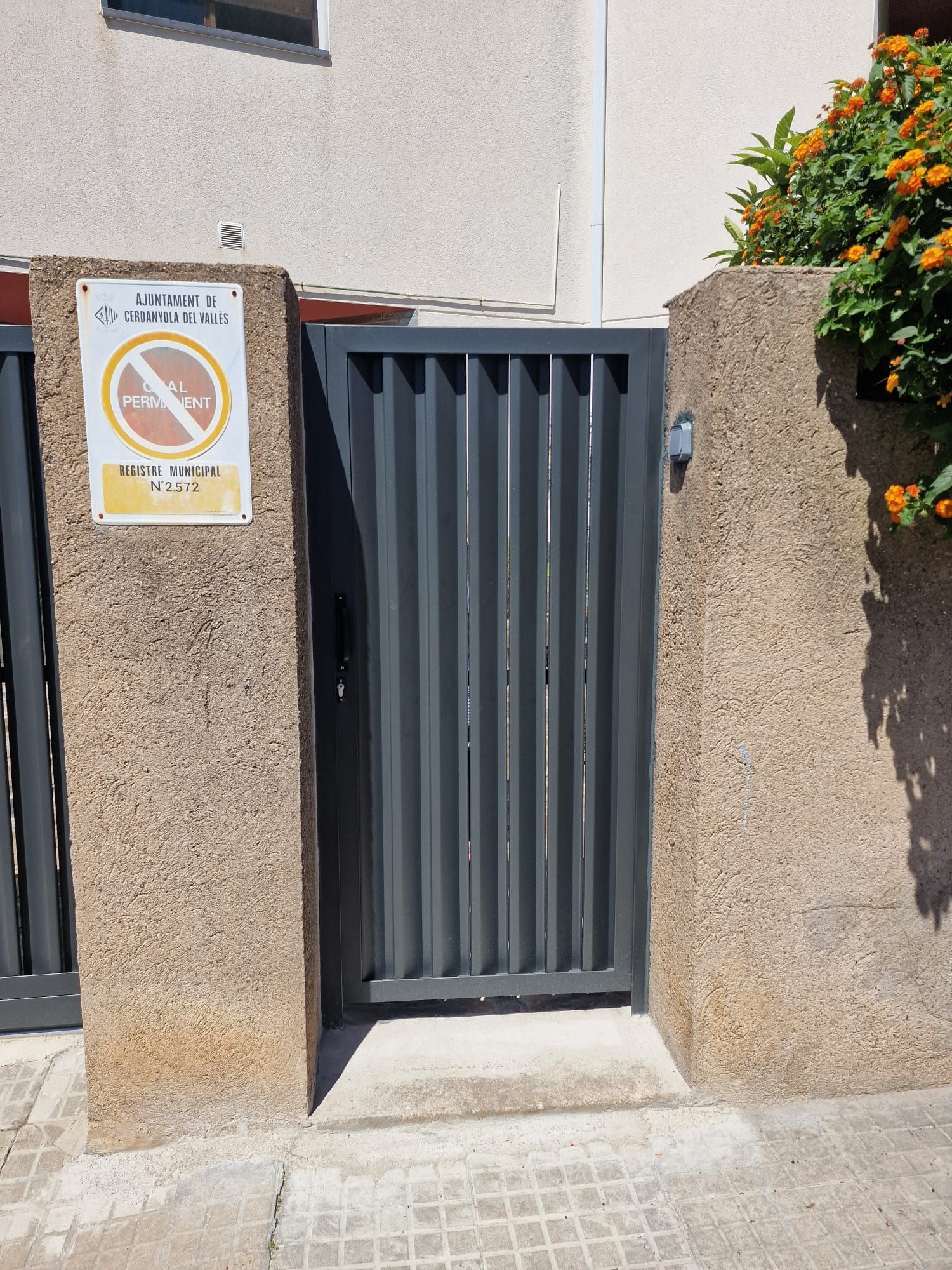 Puerta metálica gris entre muros de piedra, con un cartel de prohibido perros en el pilar izquierdo.