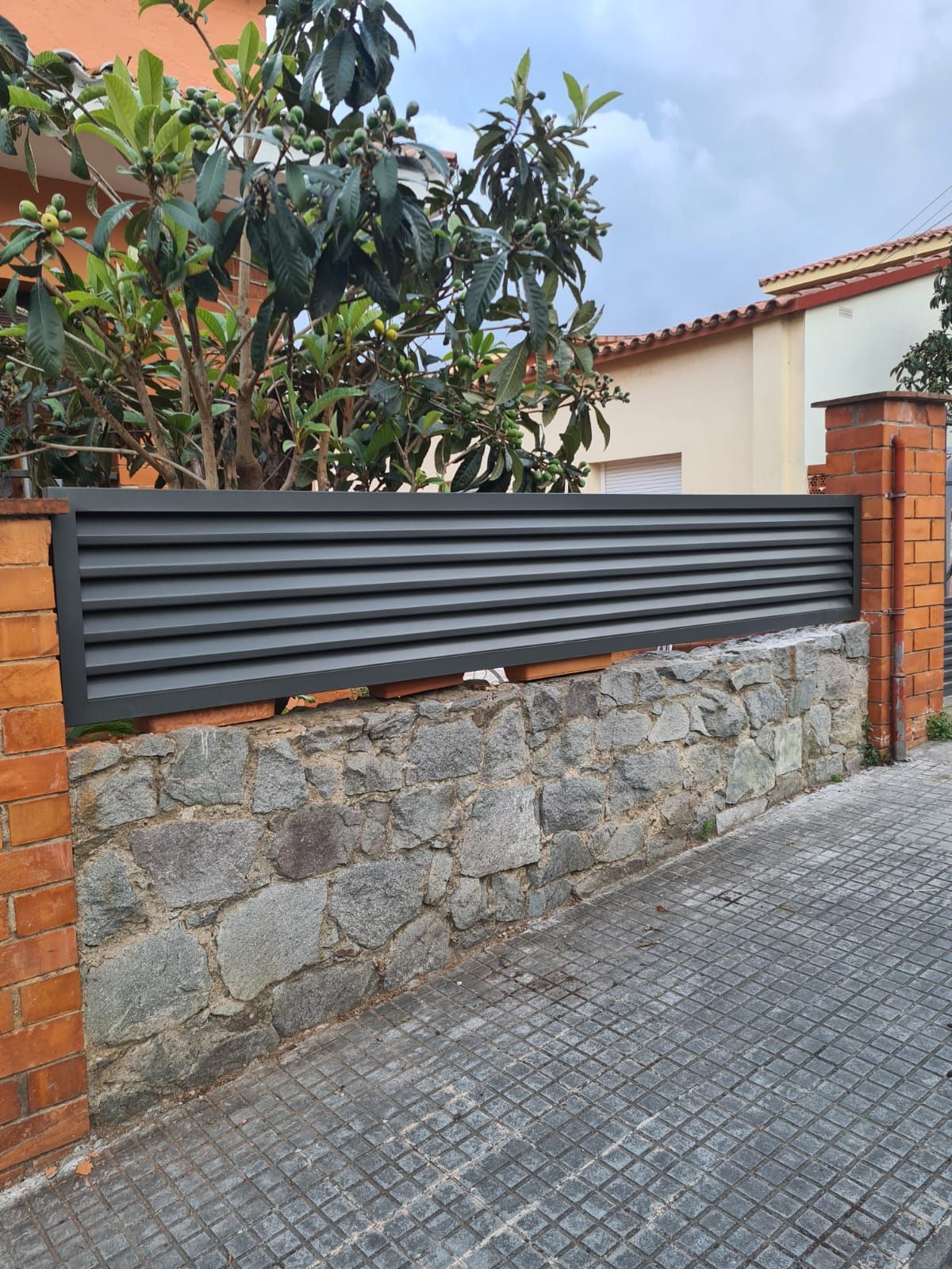 Muro de piedra y valla de metal negro junto a una entrada de vehículos, con una casa con tejado de tejas y árboles detrás