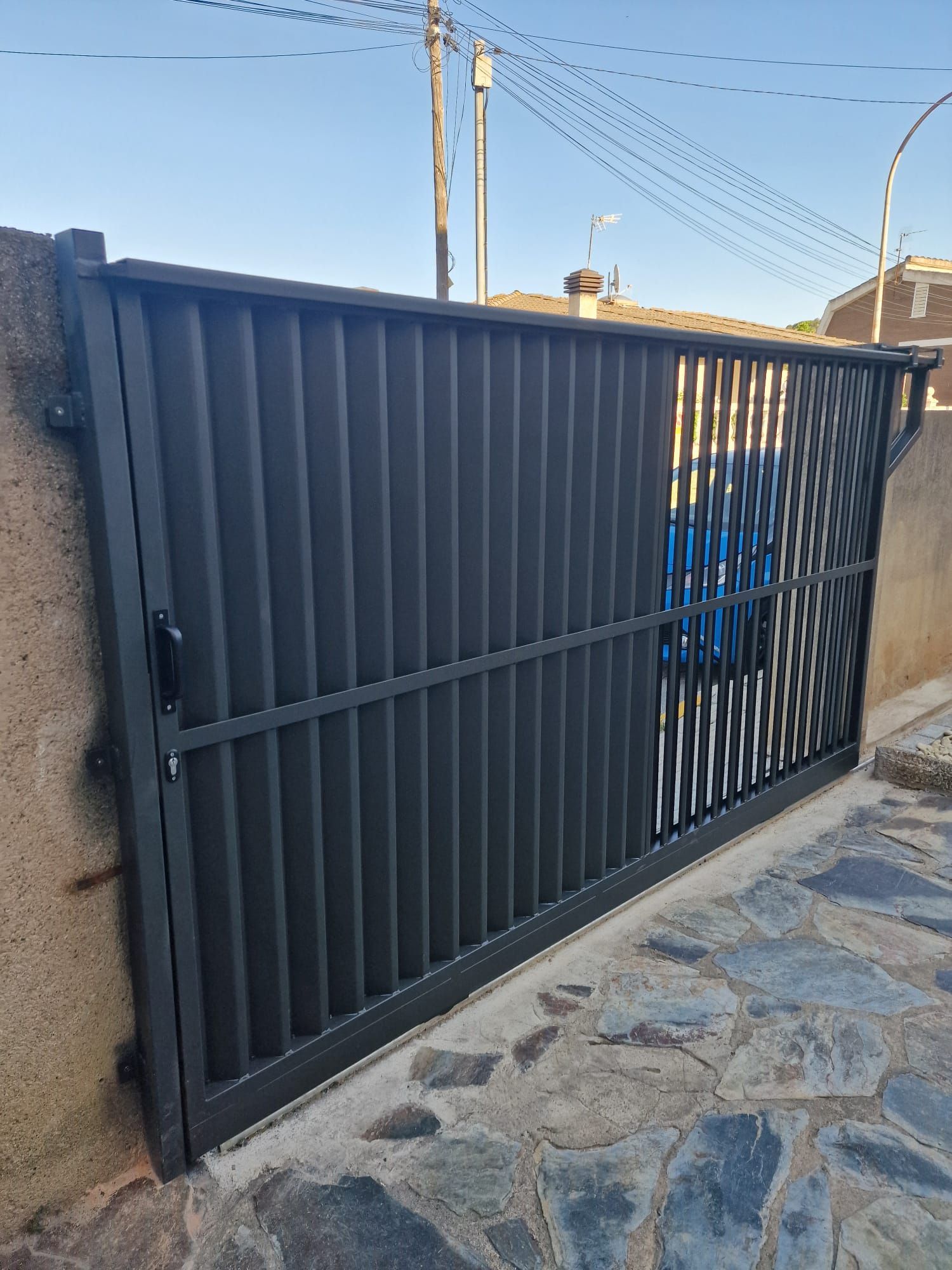 Portón corredizo de metal gris junto a un camino de piedra y un muro de estuco bajo un cielo azul