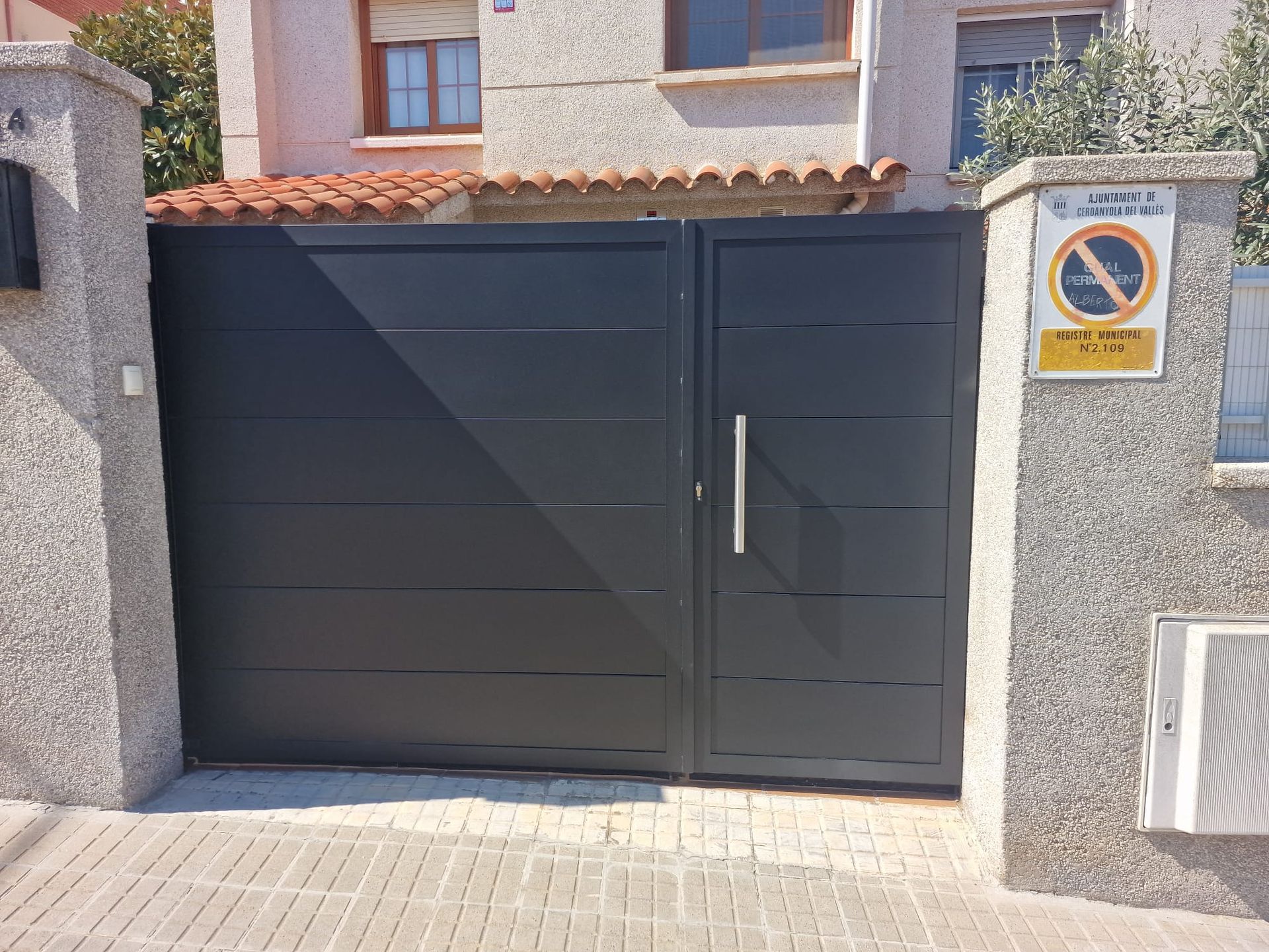 Portón de entrada de metal negro con puerta peatonal entre muros de piedra, en el exterior de una casa