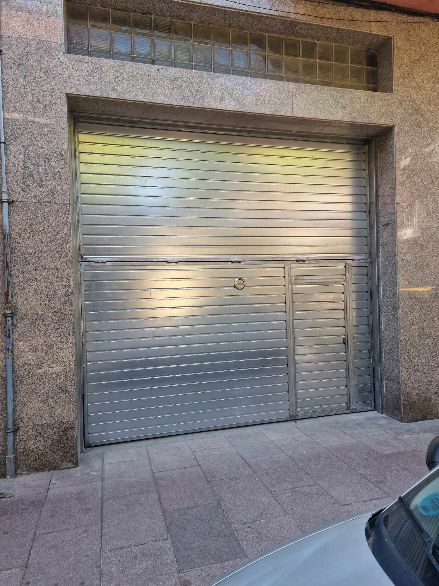 Persiana metálica plateada de un local, cerrada y con una puerta pequeña, en la fachada de piedra de un edificio.