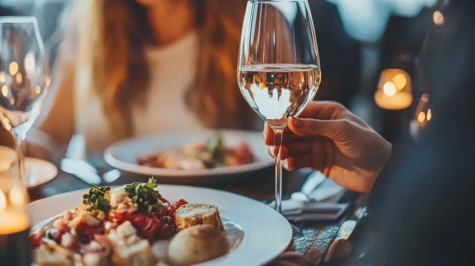 Gros plan sur une personne tenant un verre de vin à une table de restaurant avec des assiettes de nourriture et des bougies.