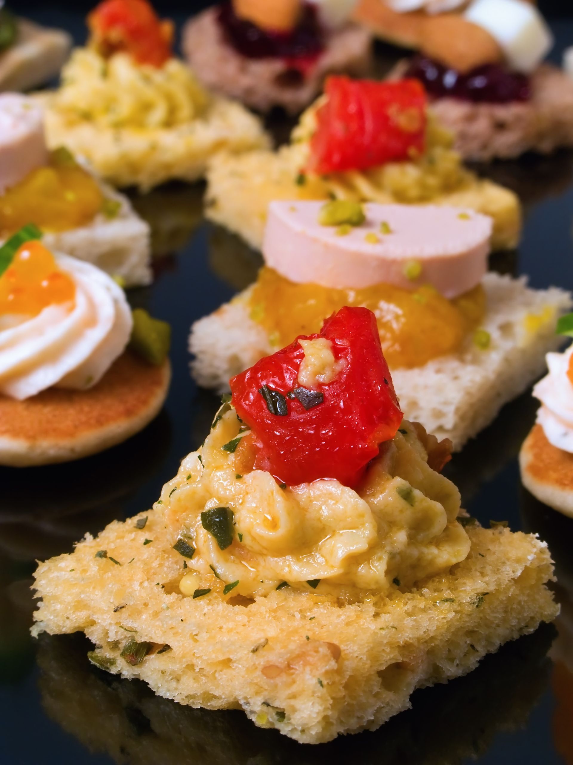 Assortiment de canapés sur une surface noire ; garnitures variées sur de petites bases de pain : crème, tomate, salade d'œufs.