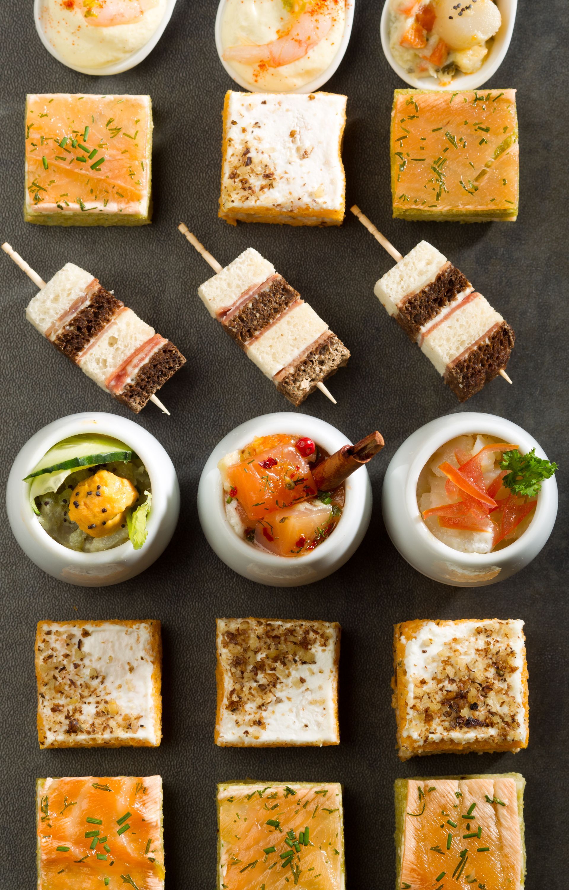 Des amuse-gueules disposés sur une surface sombre : mini-sandwichs, fruits de mer en bols, bouchées de saumon.