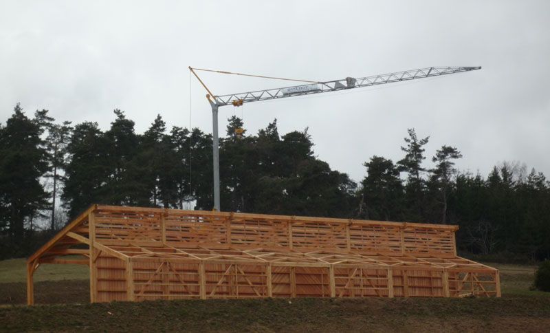Bâtiment à ossature bois en construction, avec une grue dans un champ, des arbres en arrière-plan, ciel couvert.