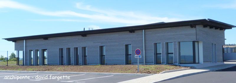 Bâtiment moderne en briques grises, avec un toit sombre et de grandes fenêtres, par une journée ensoleillée.