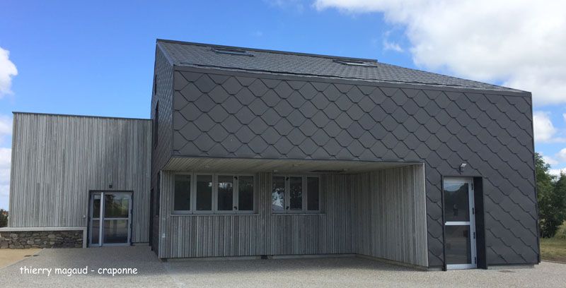 Bâtiment moderne avec toit en ardoise grise et bardage en bois, sous un ciel bleu.