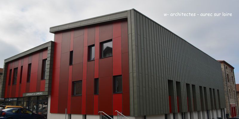 Bâtiment moderne aux panneaux rouges et gris, fenêtres rectangulaires et ciel nuageux.