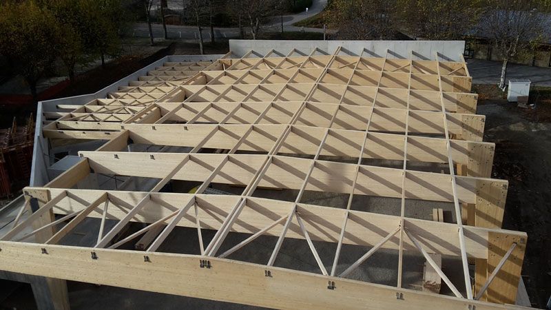 Vue aérienne d'un bâtiment en construction, montrant une charpente de toit en bois avec des poutres de soutien triangulaires.