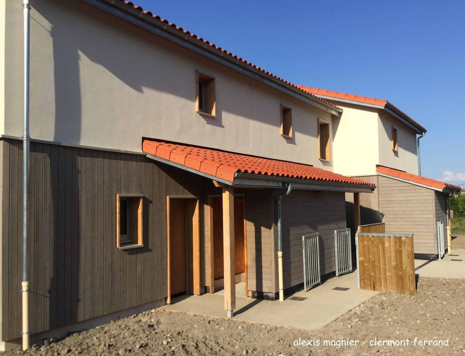 Bâtiment de deux étages avec toit de tuiles rouges, lambris et murs en stuc beige, sous un ciel bleu azur.