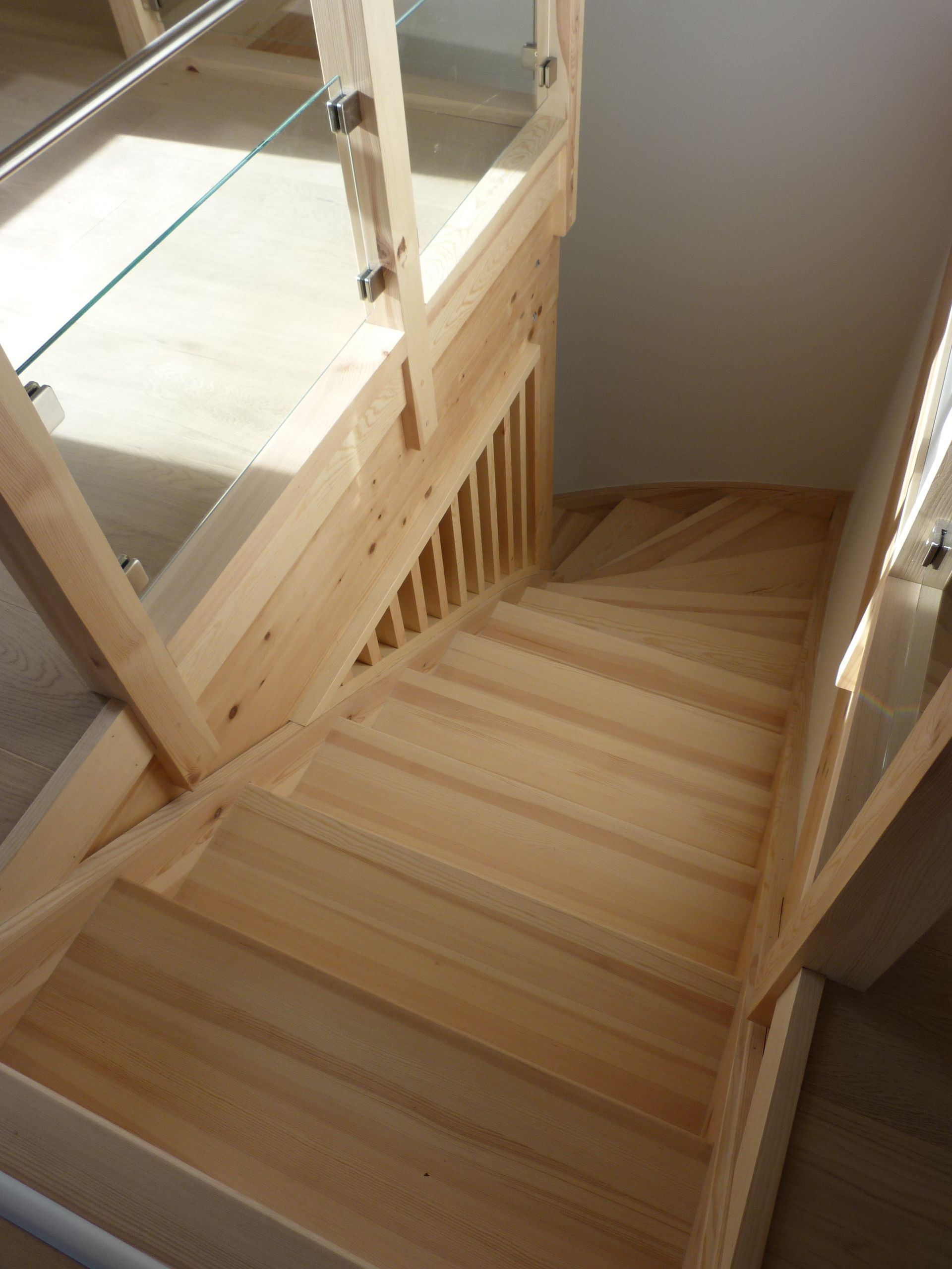 Escalier en bois avec un virage, rampe en verre et en bois, descendant dans un espace bien éclairé.