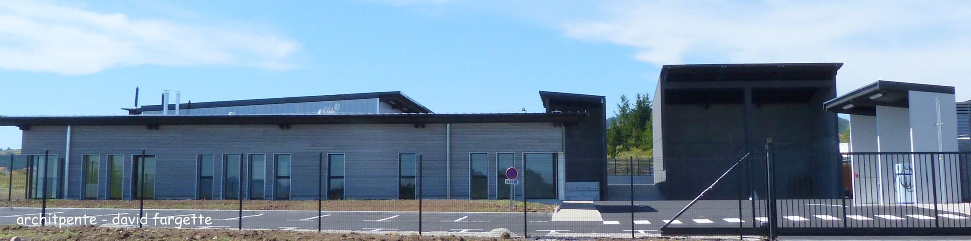 Un bâtiment gris aux grandes fenêtres et au toit plat se détachant sur un ciel bleu, visible derrière une clôture.