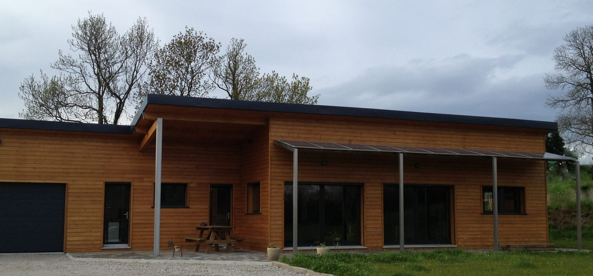 Maison en bois avec toit noir, garage et porche. Sous un ciel nuageux.
