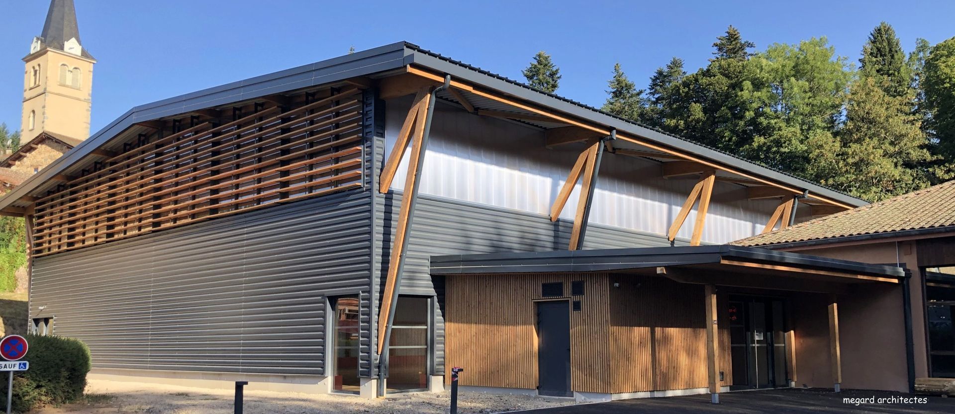 Bâtiment moderne à l'extérieur en bois et en métal, jouxtant une église à clocher.