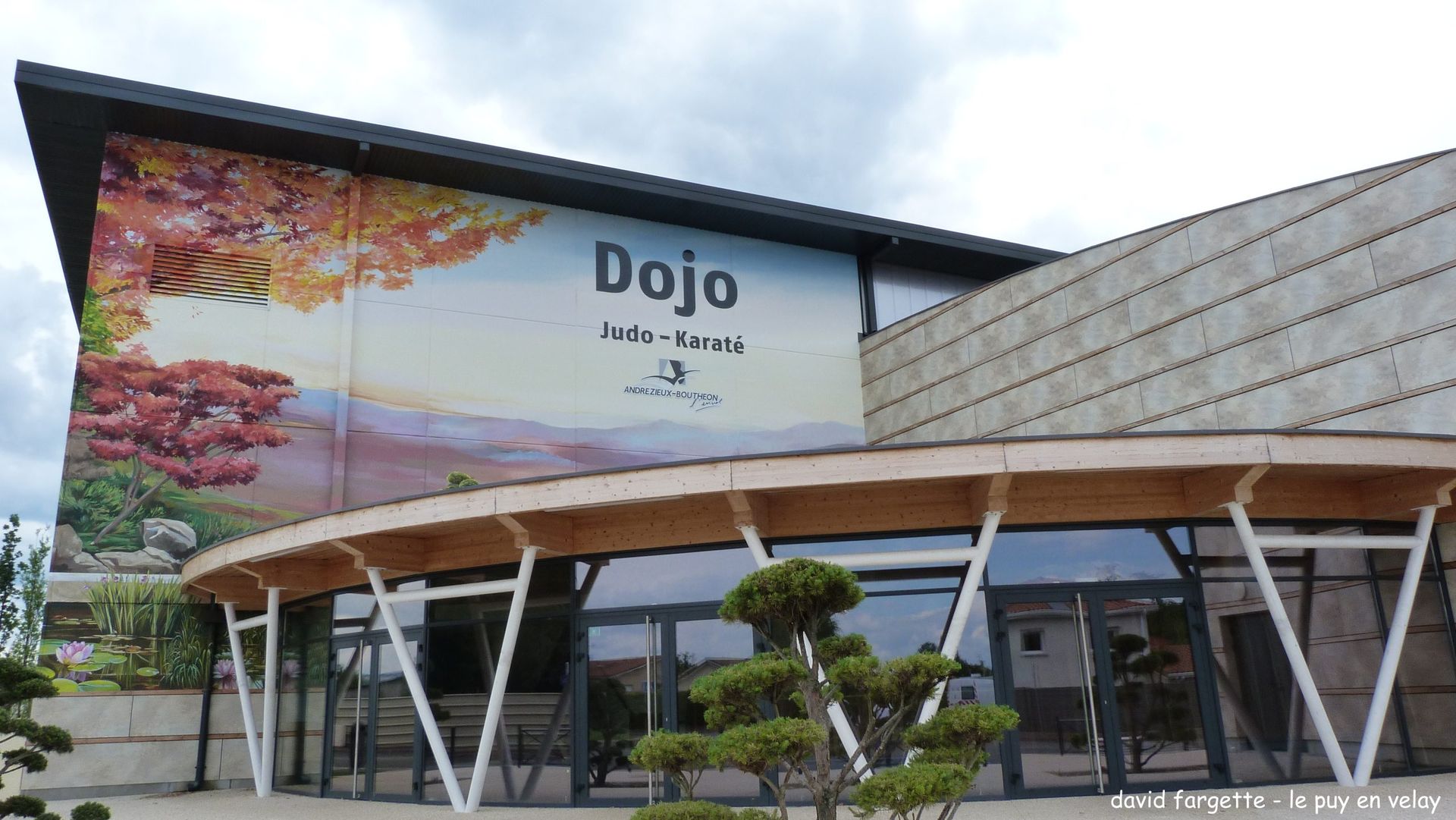 Bâtiment moderne avec enseigne Dojo, grande fresque murale, façade en verre incurvée et jardin paysager.
