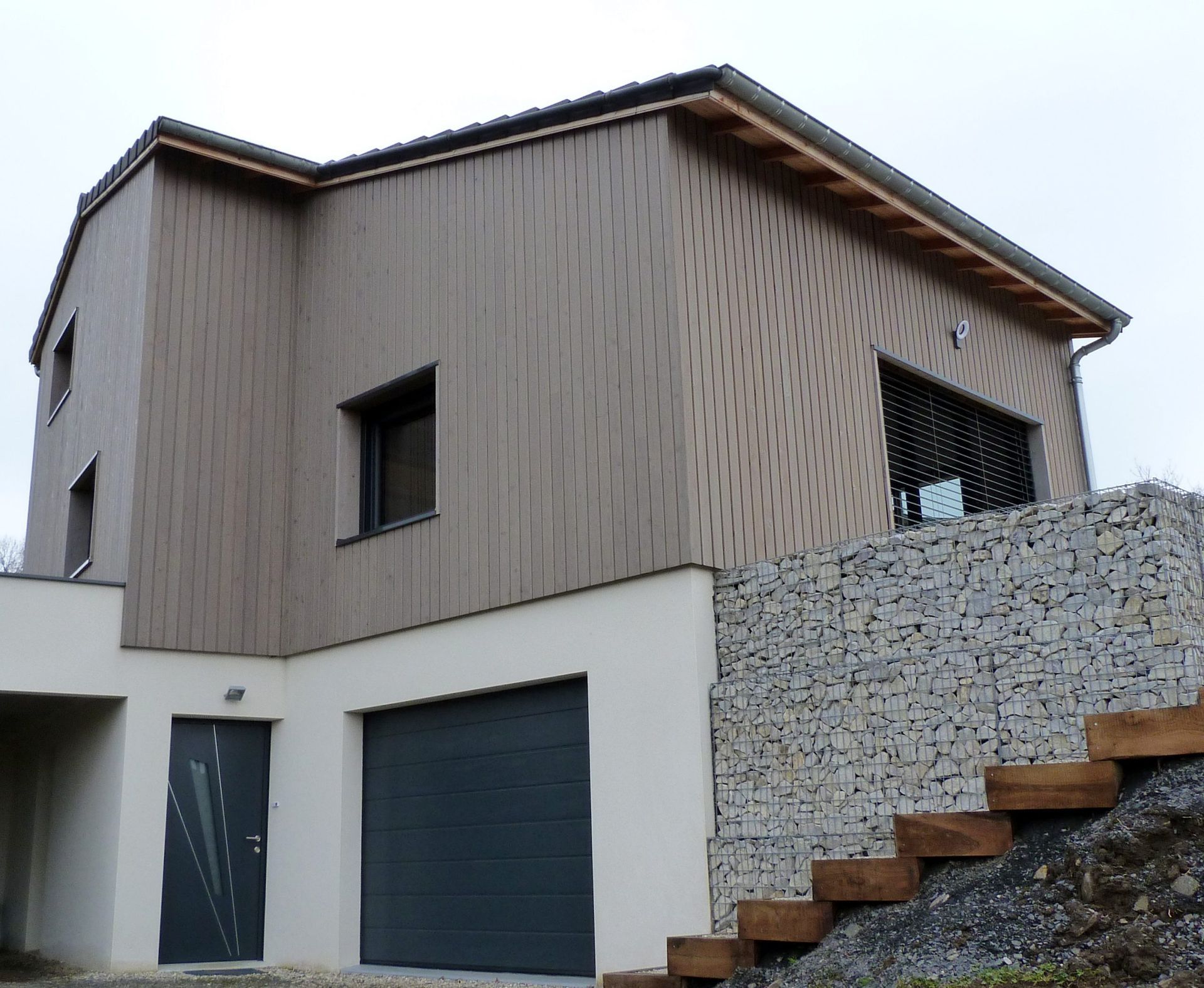 Maison moderne à deux étages avec bardage beige, garage et mur en pierre avec marches en bois.