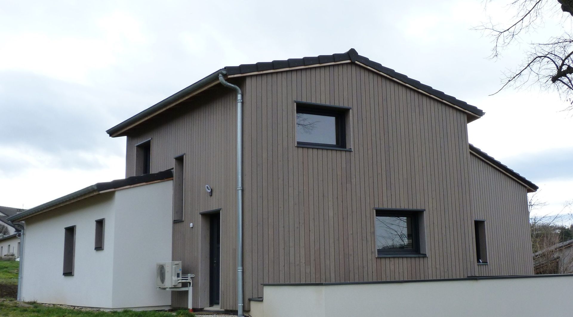 Maison moderne à deux étages avec bardage vertical brun, murs blancs et encadrements de fenêtres foncés, sous un ciel nuageux.