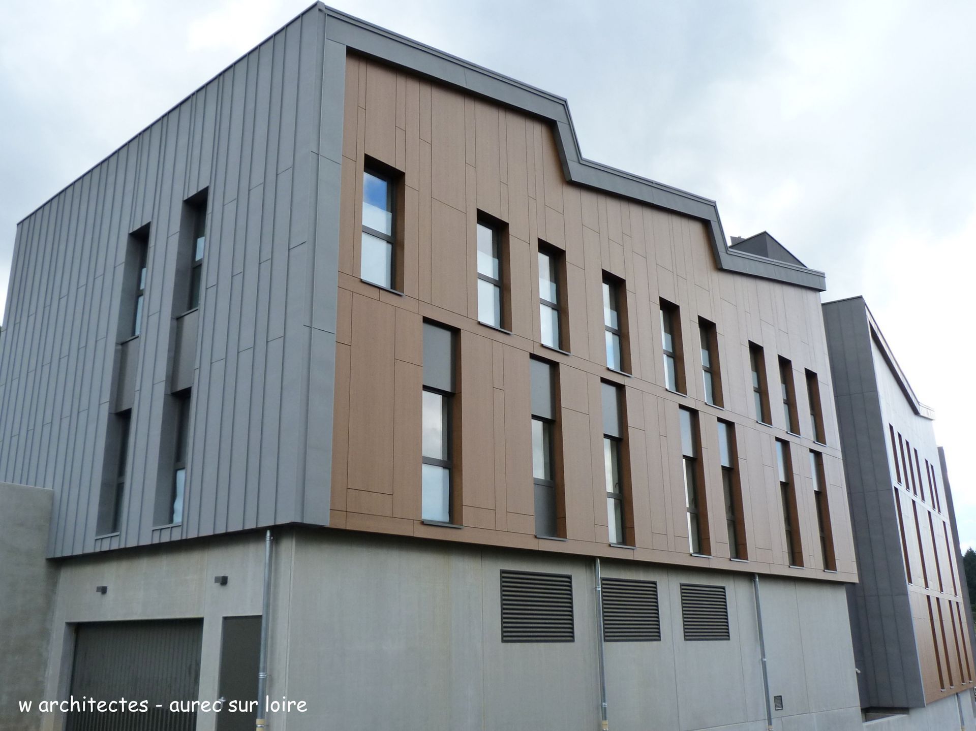 Bâtiment moderne avec une façade brune et grise, des fenêtres rectangulaires et une base en béton.