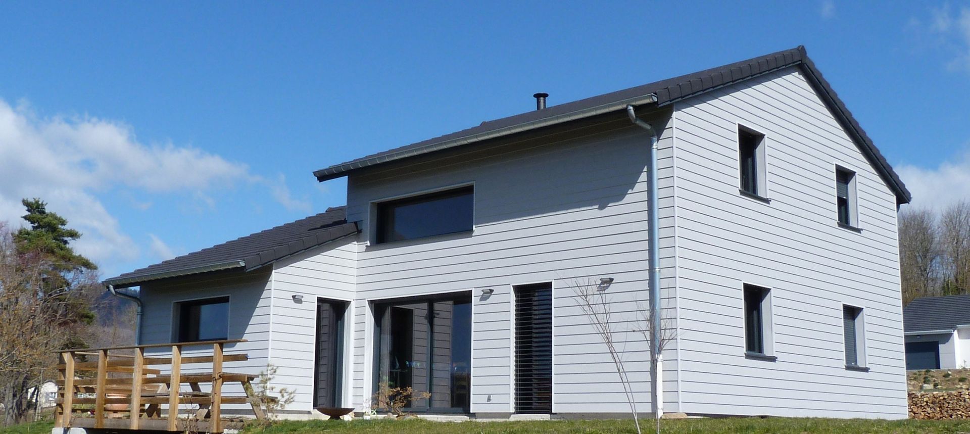Maison moderne à deux étages avec bardage gris, toit et fenêtres sombres, se détachant sur un ciel bleu.