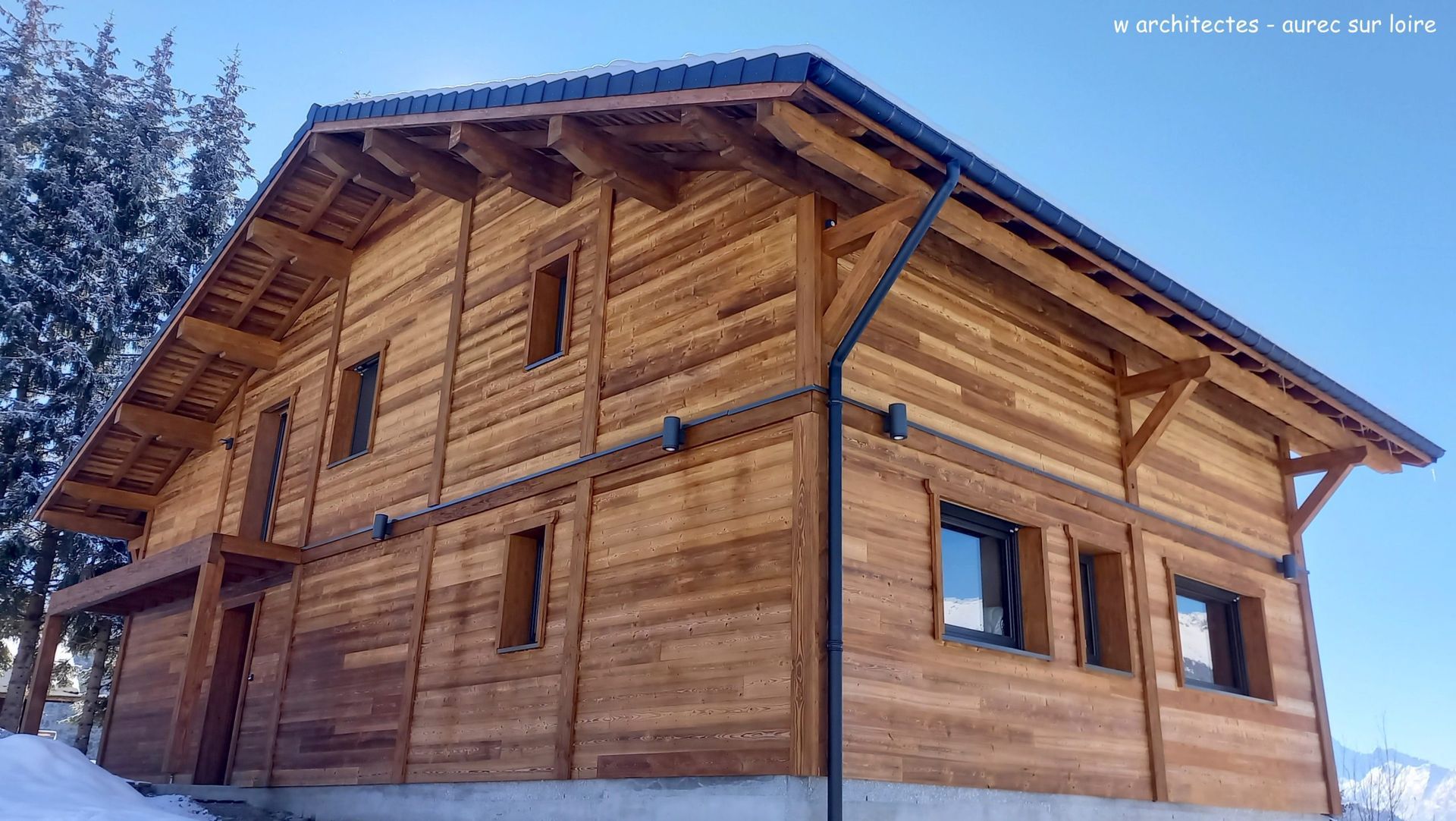 Chalet en bois au toit et aux fenêtres sombres, situé dans un paysage enneigé et ensoleillé.