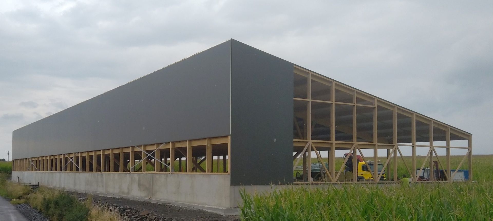 Construction d'une grande grange aux murs gris, avec une charpente en bois apparente et une base en béton, dans un champ herbeux.