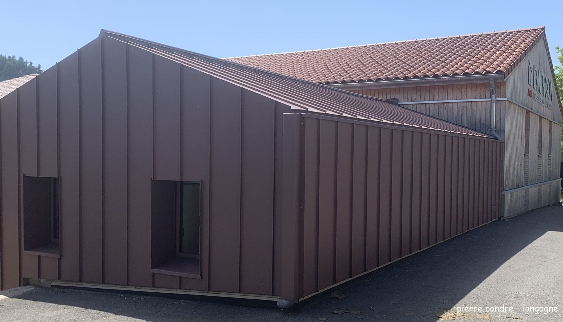 Bâtiment revêtu de métal brun avec deux fenêtres rectangulaires, relié à une structure en bois avec un toit de tuiles rouges.