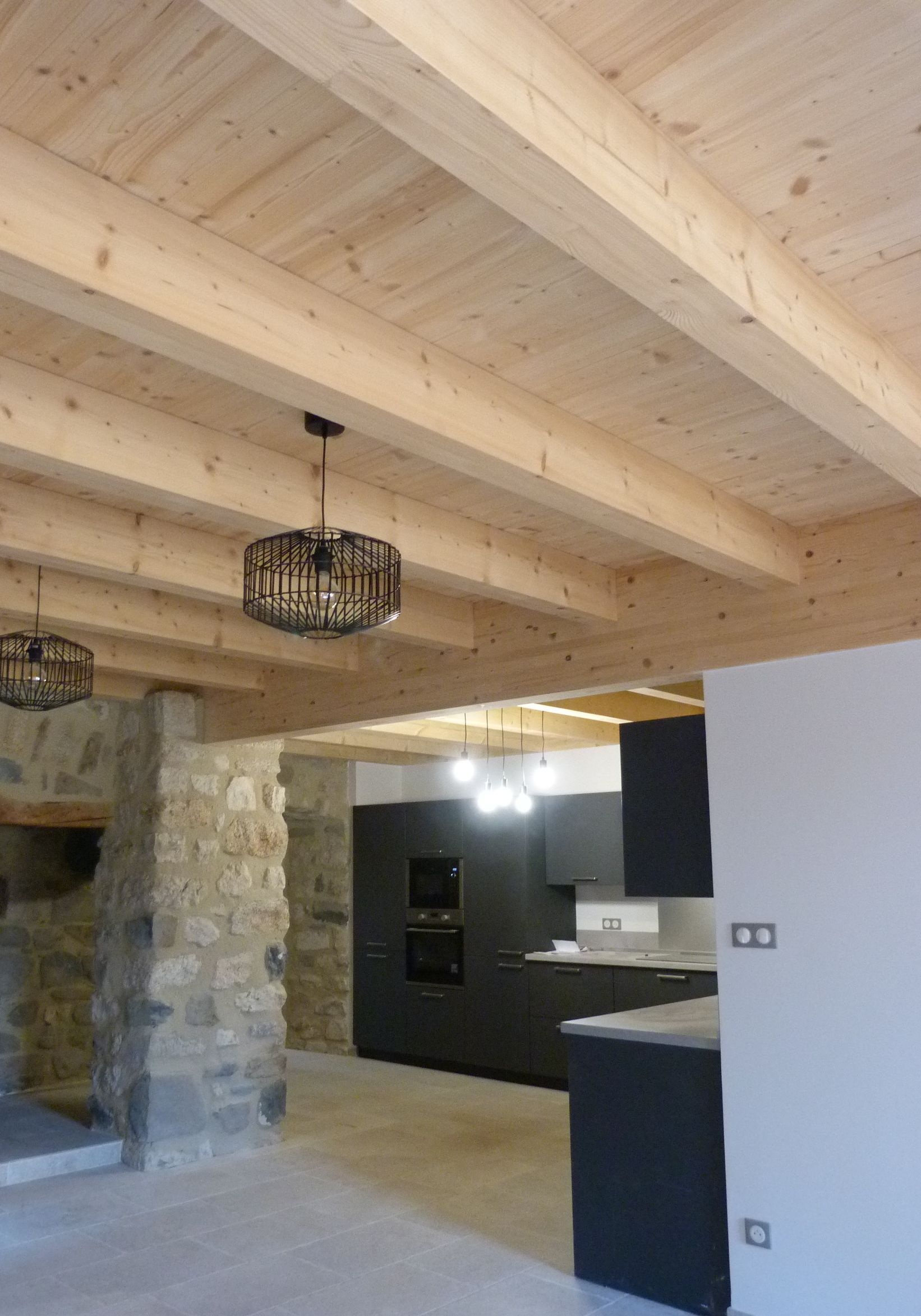 Plafond à poutres apparentes, mur en pierre, cuisine sombre, lustres modernes.