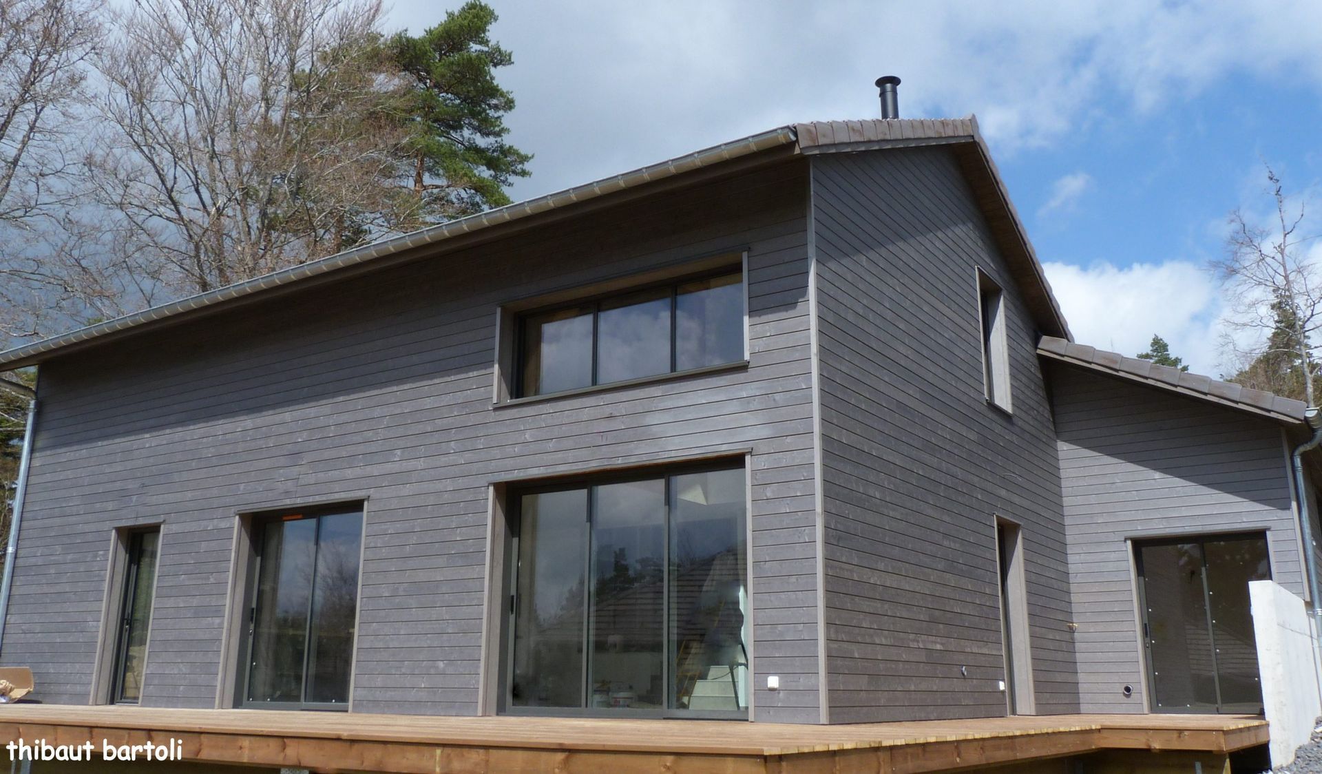 Maison à deux étages avec un revêtement extérieur gris foncé, de grandes fenêtres et une terrasse en bois.
