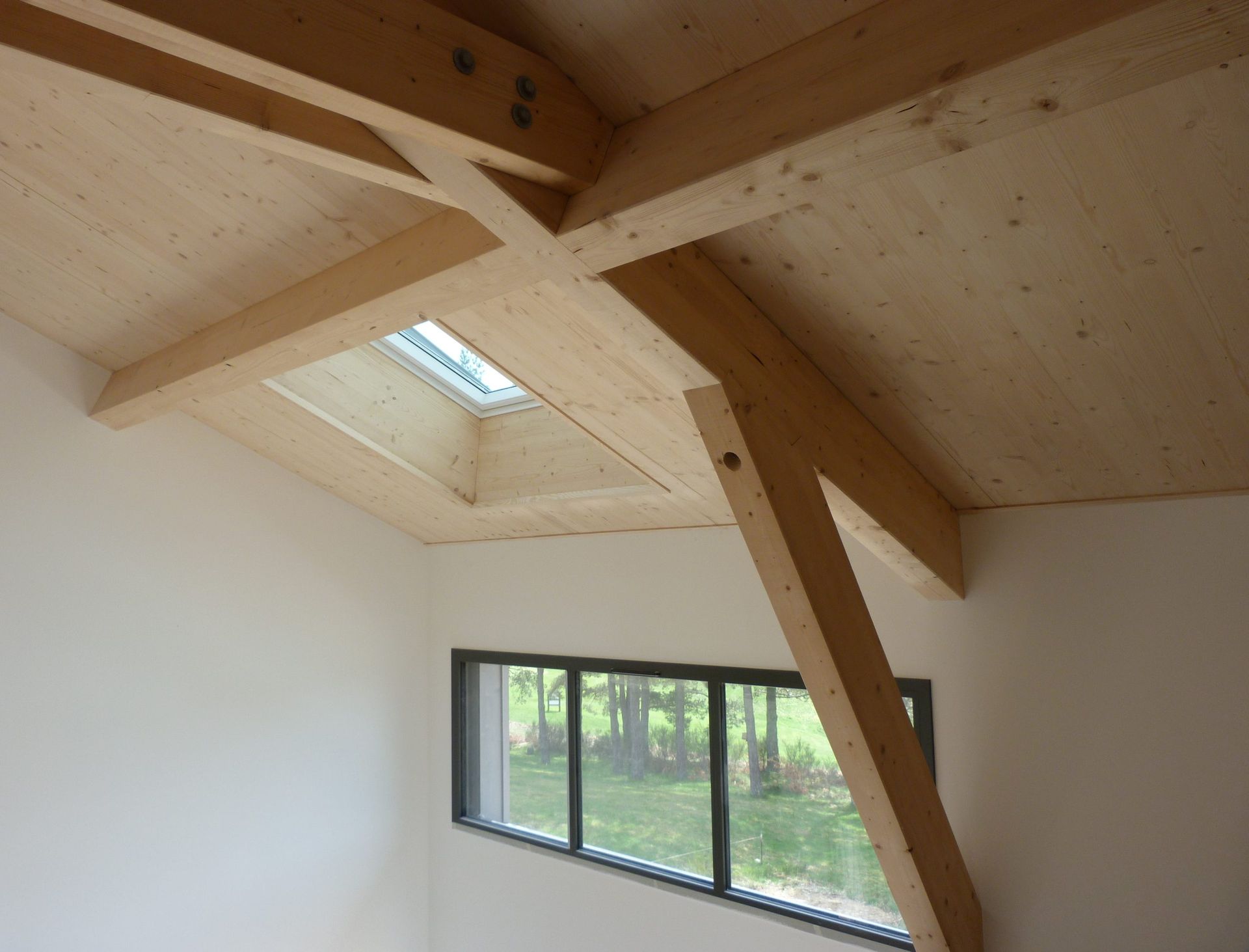 Vue intérieure d'une pièce moderne avec poutres apparentes et puits de lumière au-dessus d'une fenêtre donnant sur les arbres.