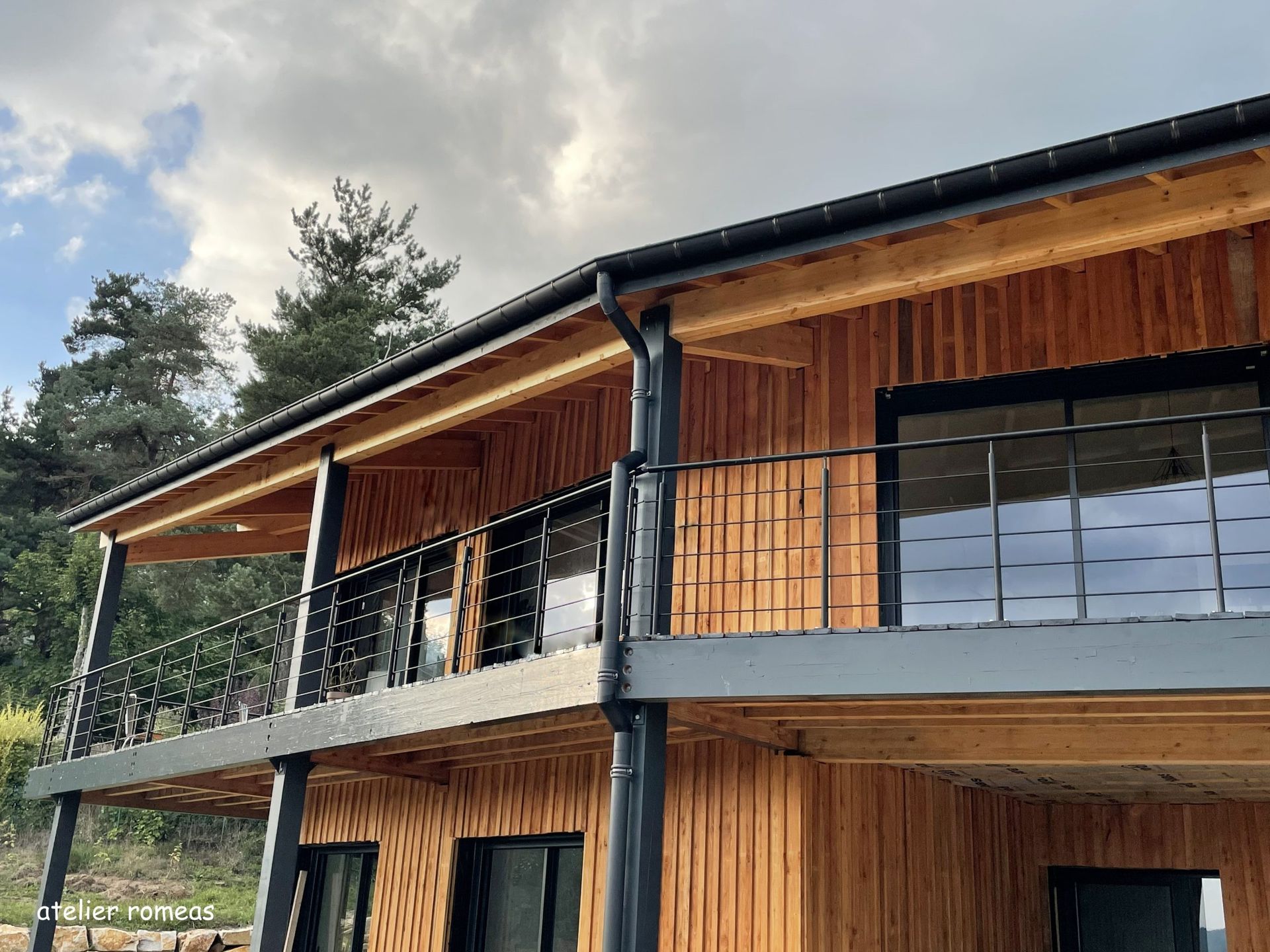 Bâtiment en bois de deux étages avec des balustrades et des boiseries noires, se détachant sur un ciel nuageux.