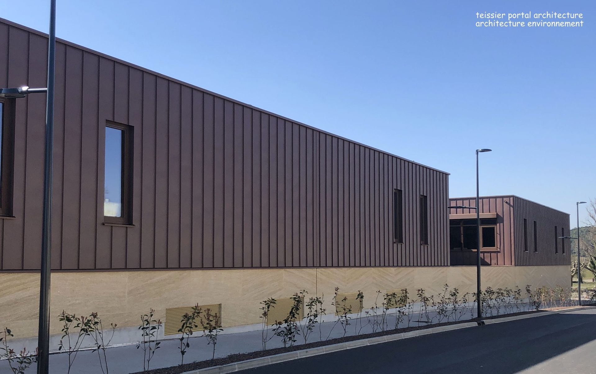 Un bâtiment revêtu de métal brun, percé de petites fenêtres rectangulaires, surplombe un mur de pierre beige. Le ciel est d'un bleu limpide.
