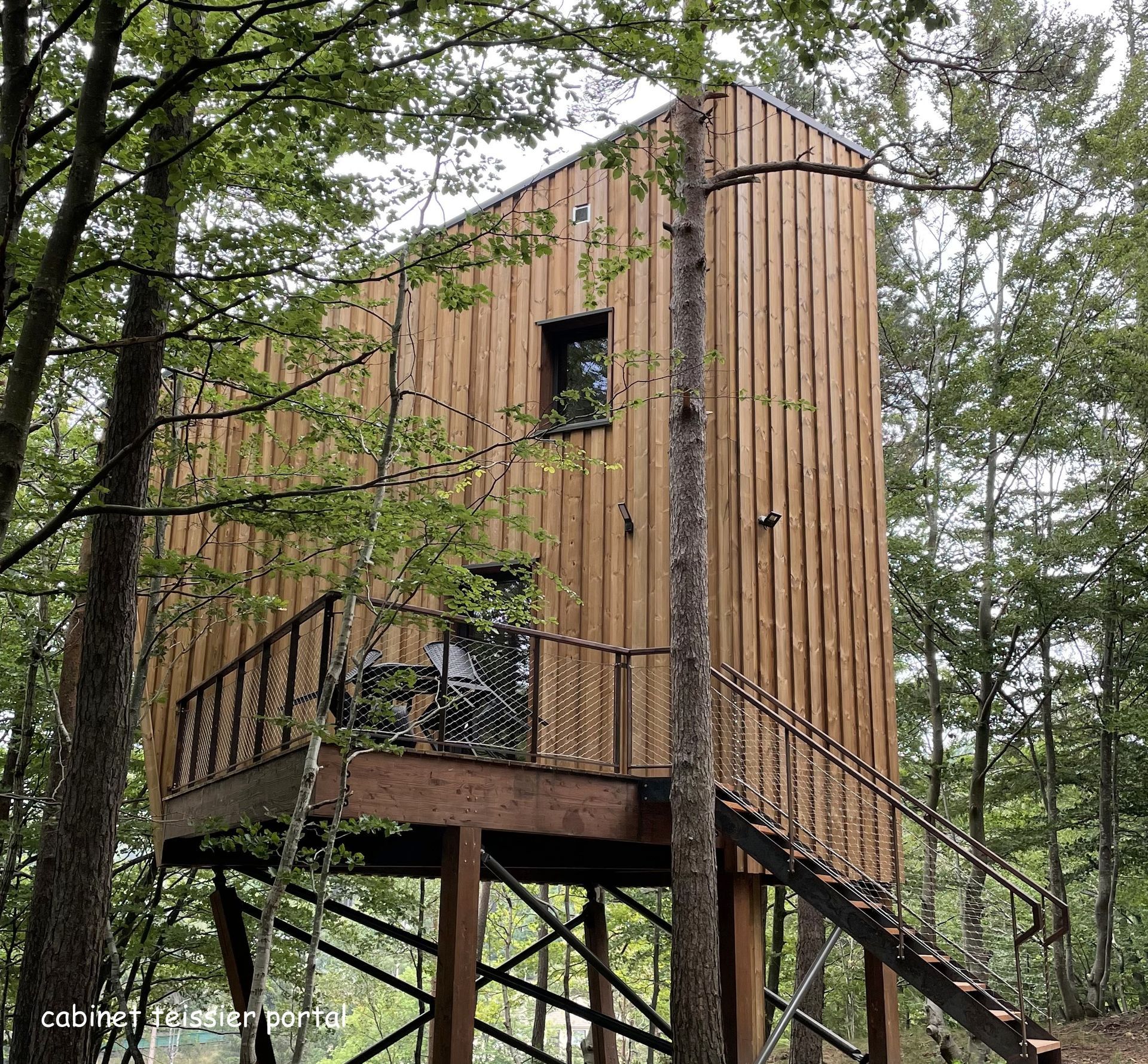 Cabane en bois sur pilotis dans une forêt. Elle possède un petit balcon et un escalier menant à l'étage.