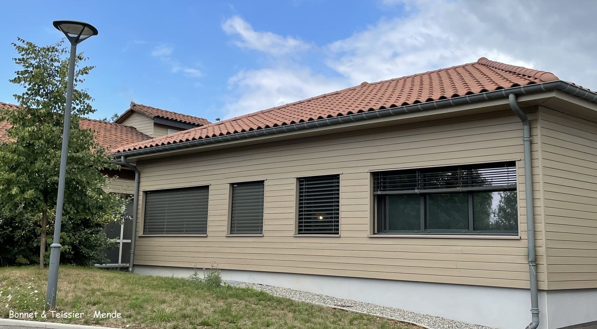 Bâtiment au revêtement brun clair, au toit de tuiles en terre cuite et aux nombreuses fenêtres ; un lampadaire se trouve au premier plan.