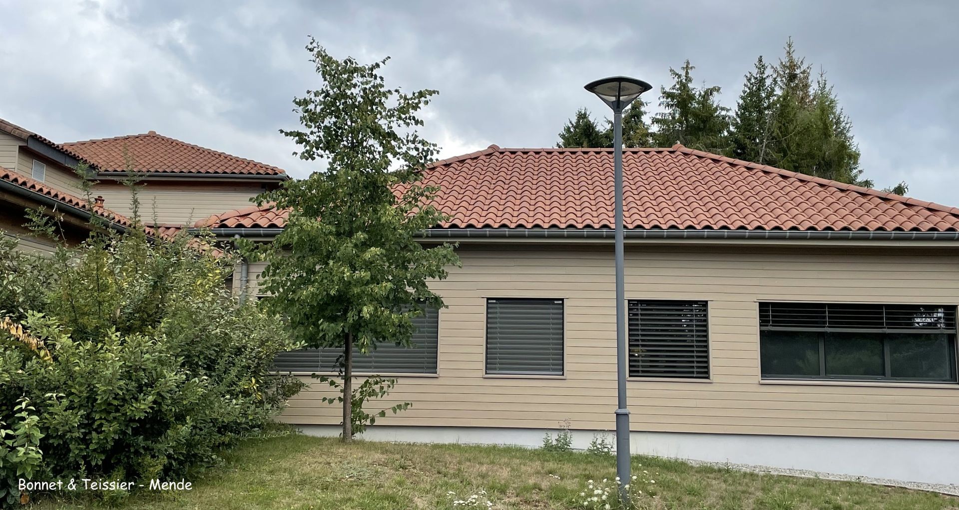 Bâtiment beige au toit de tuiles orange ; fenêtres condamnées ; petit arbre devant ; ciel nuageux.