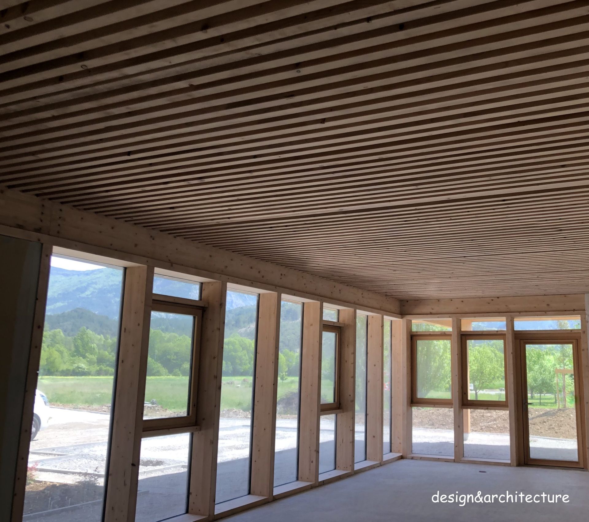 Des poutres et des lattes de bois forment le plafond et les encadrements de fenêtres d'une pièce. De grandes fenêtres offrent une vue sur le paysage extérieur.
