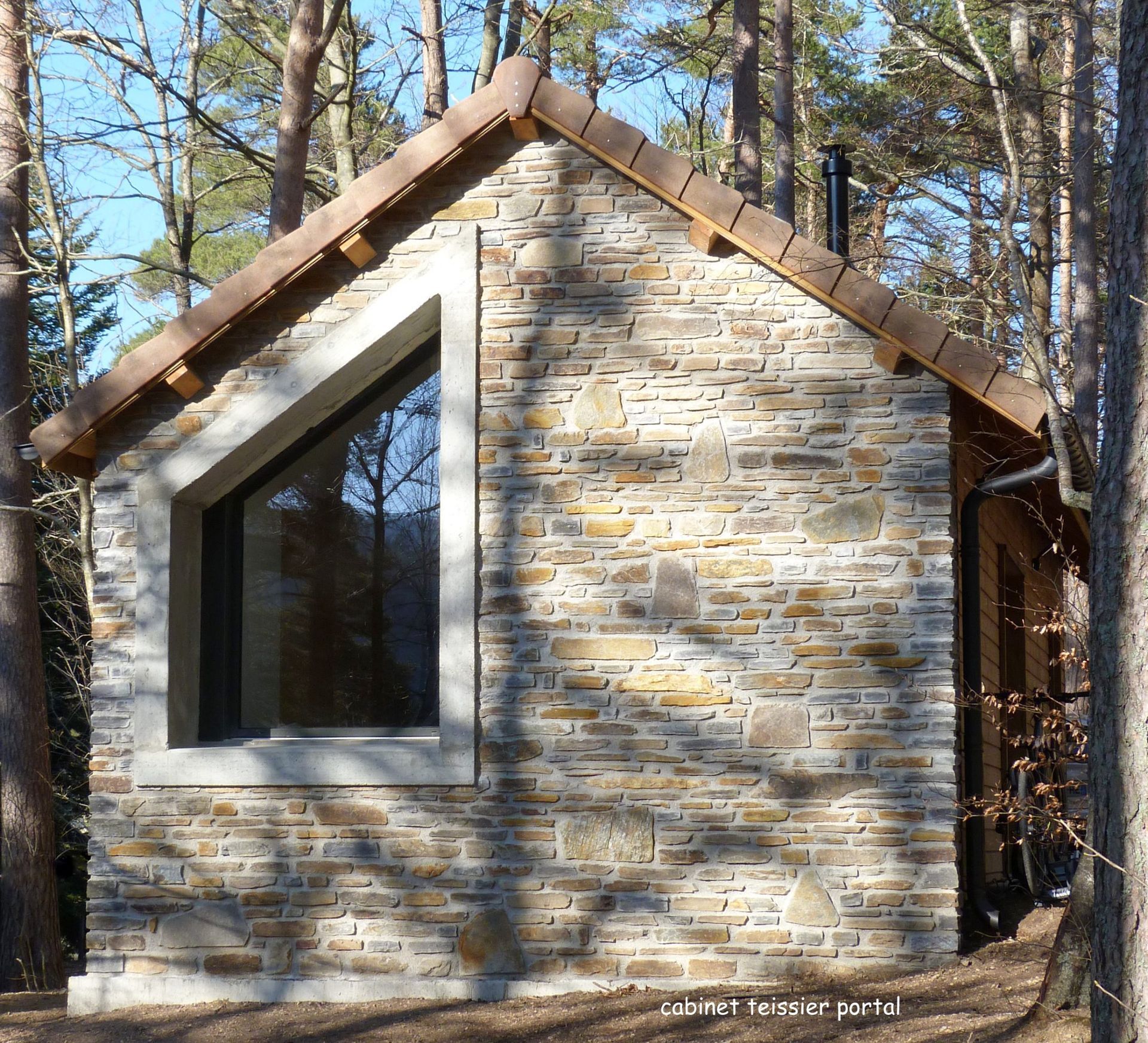 Cabane en pierre avec une grande fenêtre en biais, toit de tuiles brunes, située en forêt.