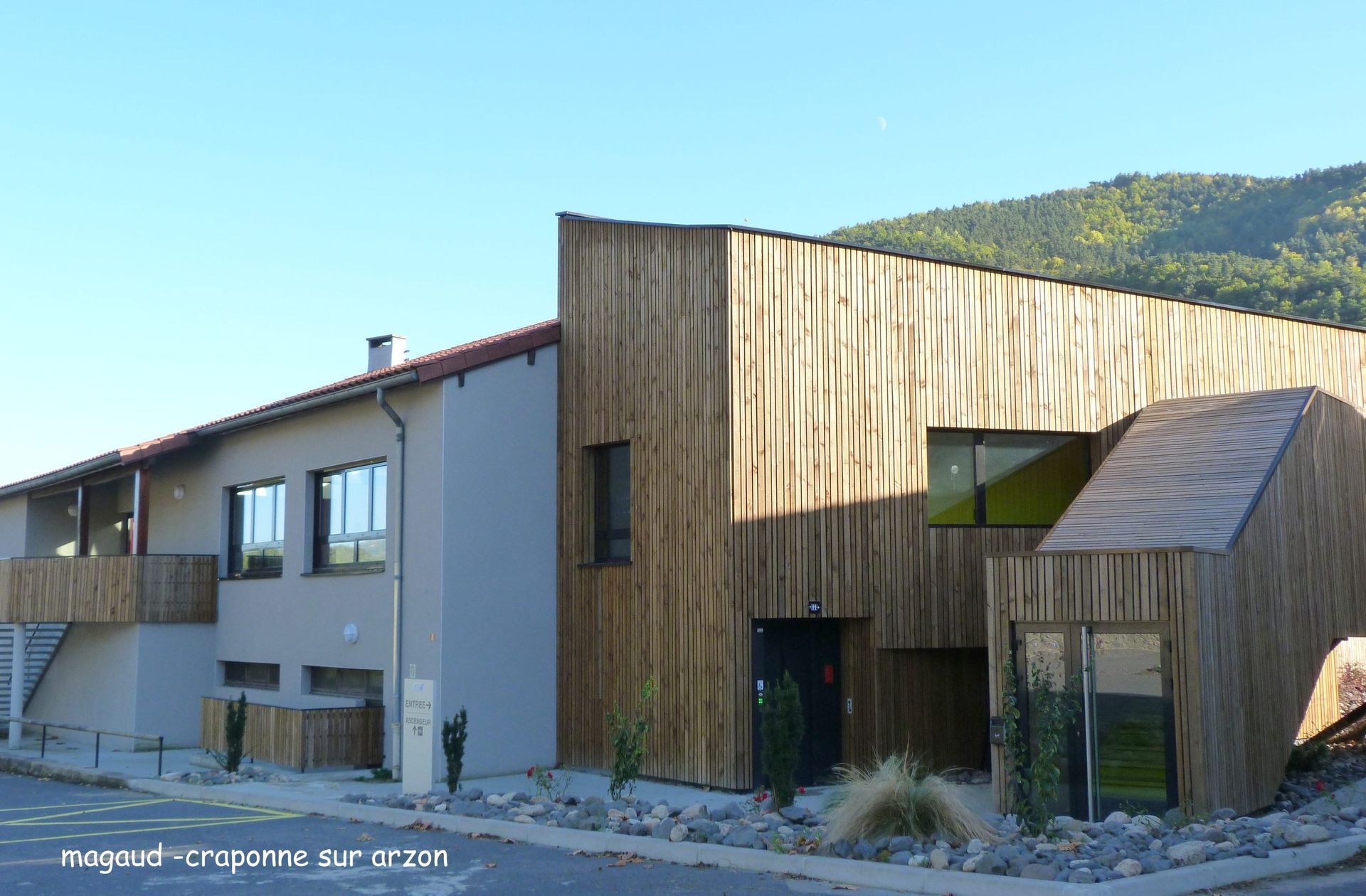 Bâtiment moderne avec bardage en bois et façade en stuc gris. Montagnes en arrière-plan.