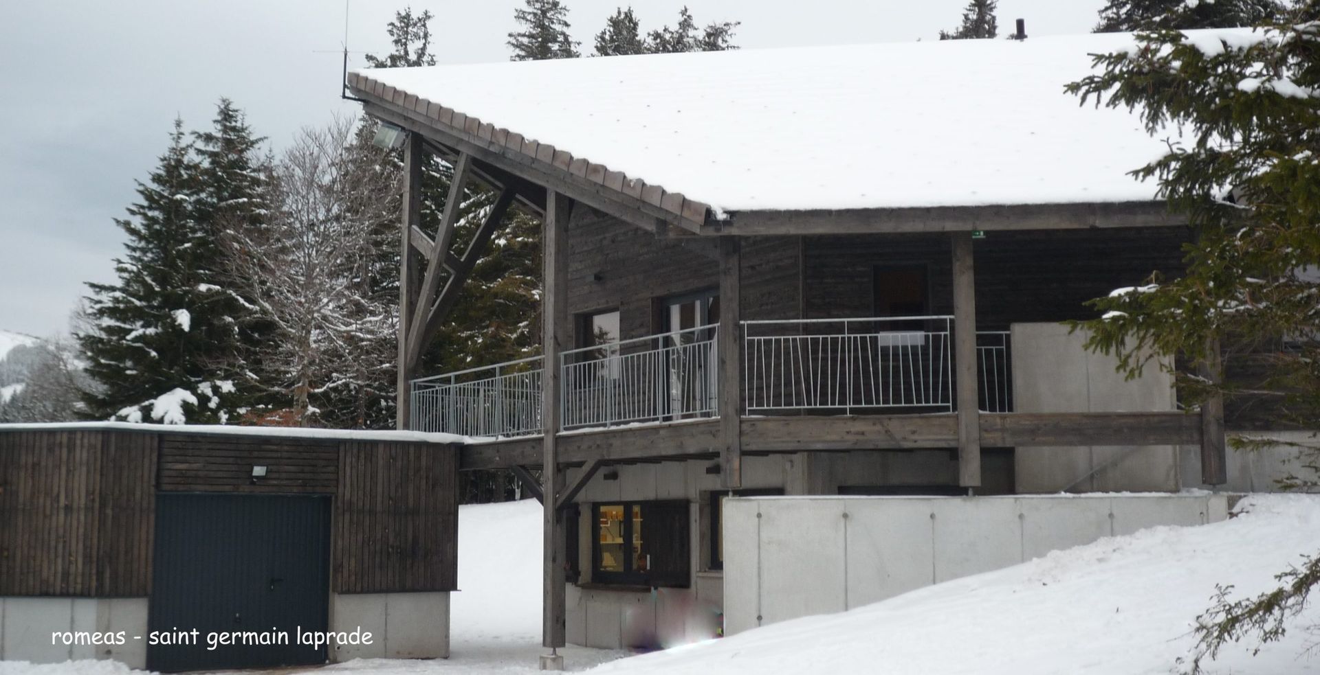 Chalet enneigé avec bardage en bois et balcon, arbres en arrière-plan.