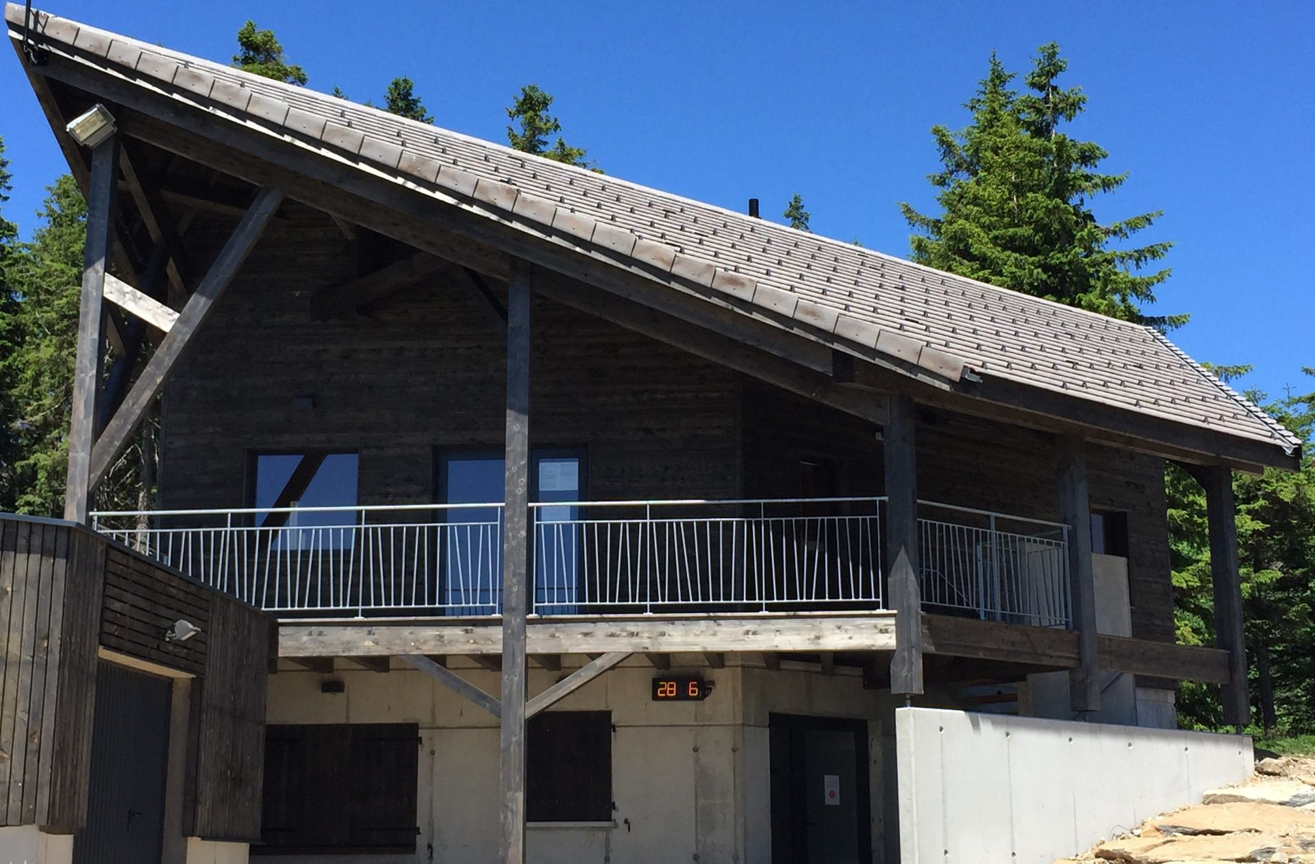 Cabane en bois avec balcon, toit gris, se détachant sur un ciel bleu et des arbres.