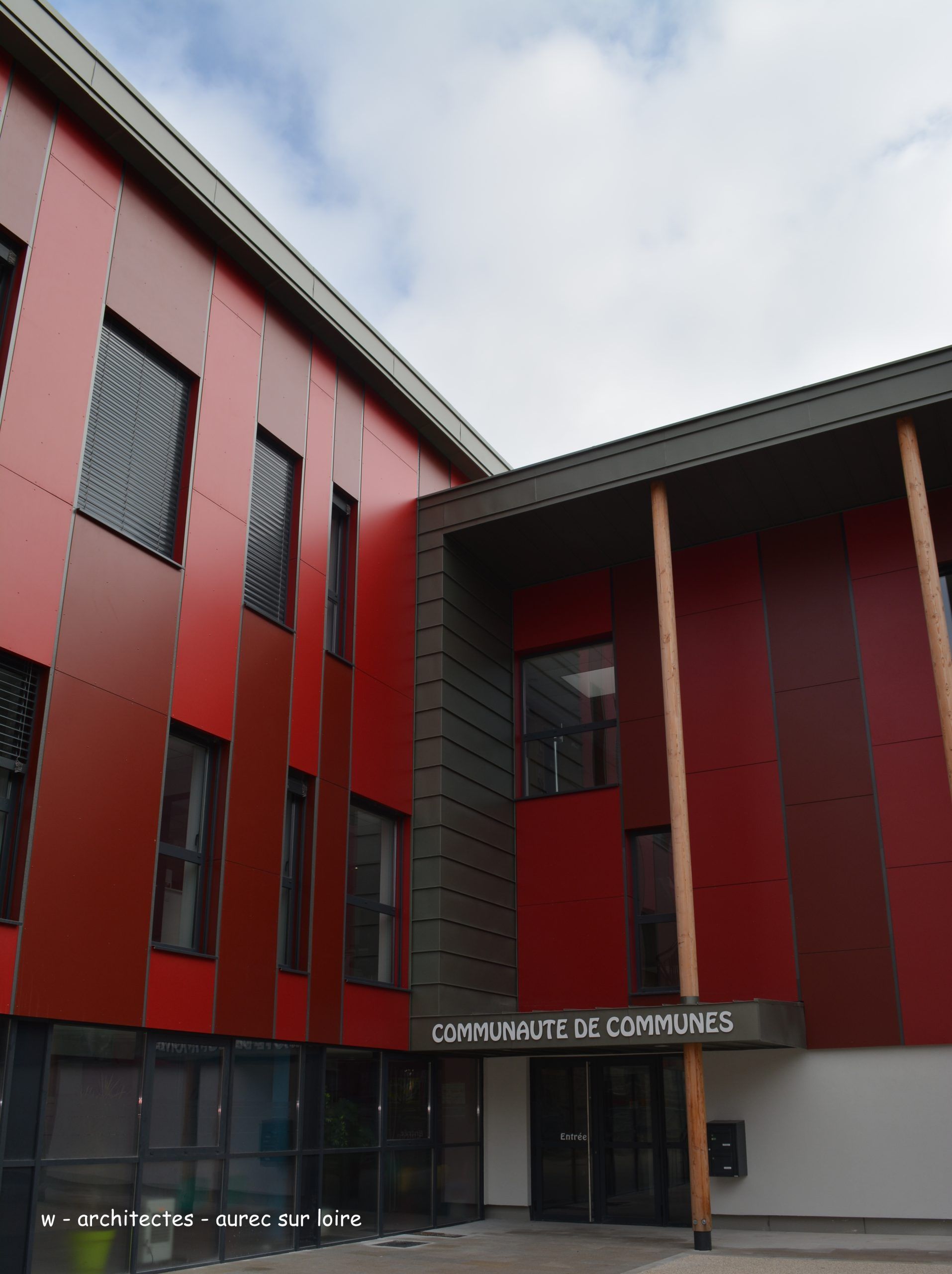 Bâtiment moderne rouge et gris, panneau « Centre Interco de Communes » au-dessus de l'entrée, ciel bleu.