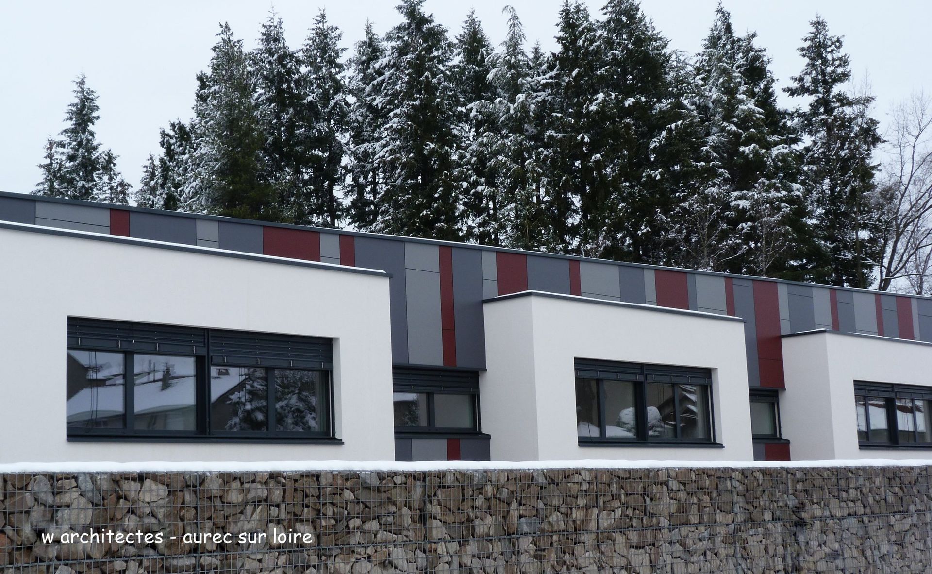 Bâtiment blanc avec des touches de rouge et de gris, des fenêtres sombres et un mur de pierre devant des arbres.