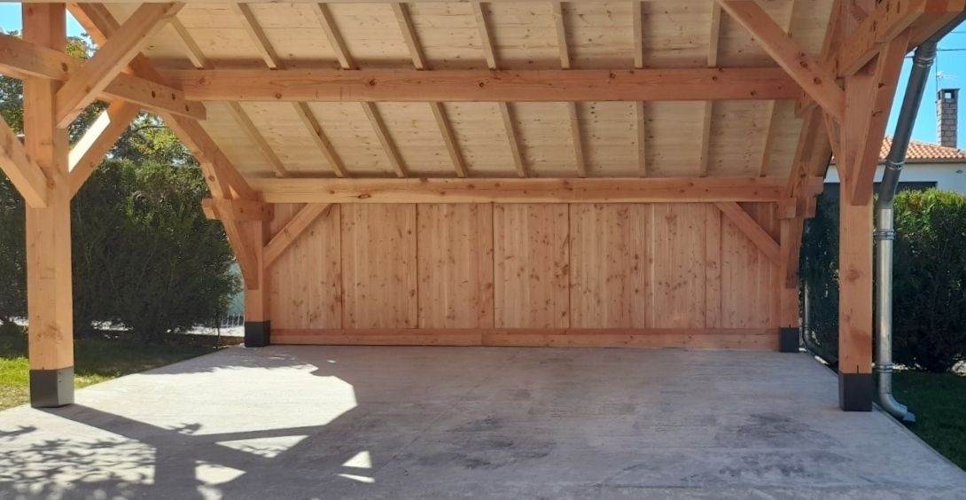 Abri voiture en bois avec sol en béton, construit avec des poutres apparentes et des lambris, devant un espace vert.