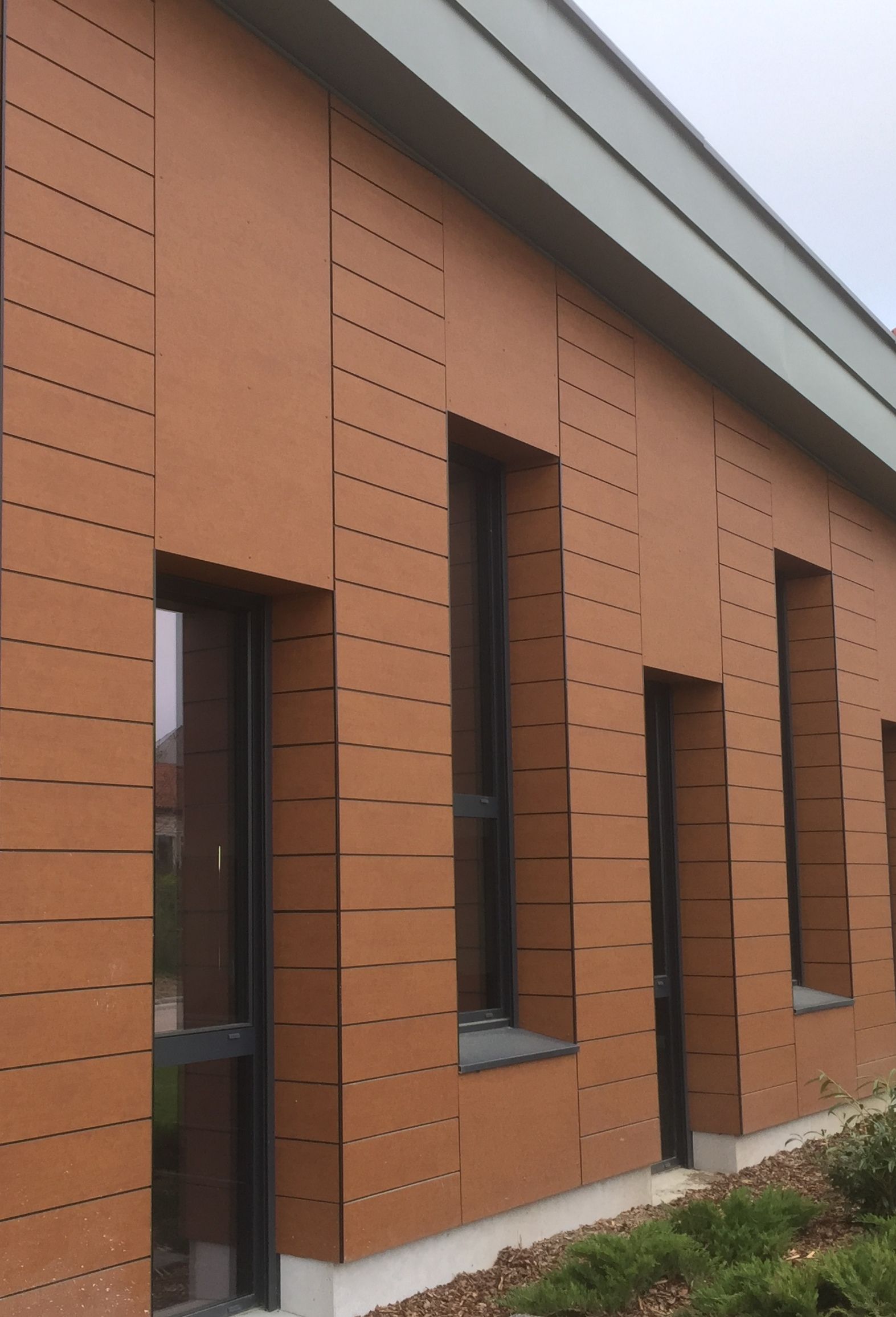 Façade d'un bâtiment brun avec panneaux verticaux, fenêtres et toit vert.