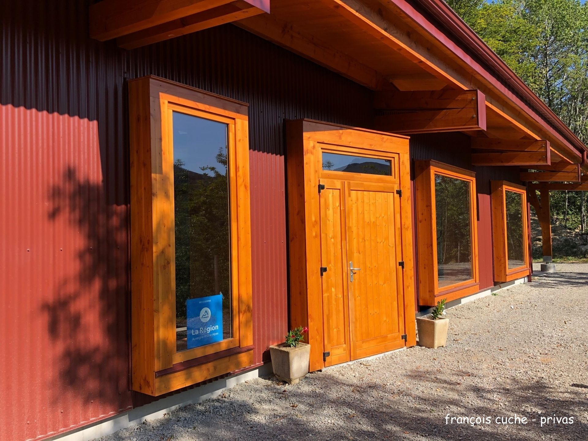 Bâtiment rouge avec fenêtres et porte à encadrement en bois. Sol de gravier, plantes et vue partielle sur le ciel.