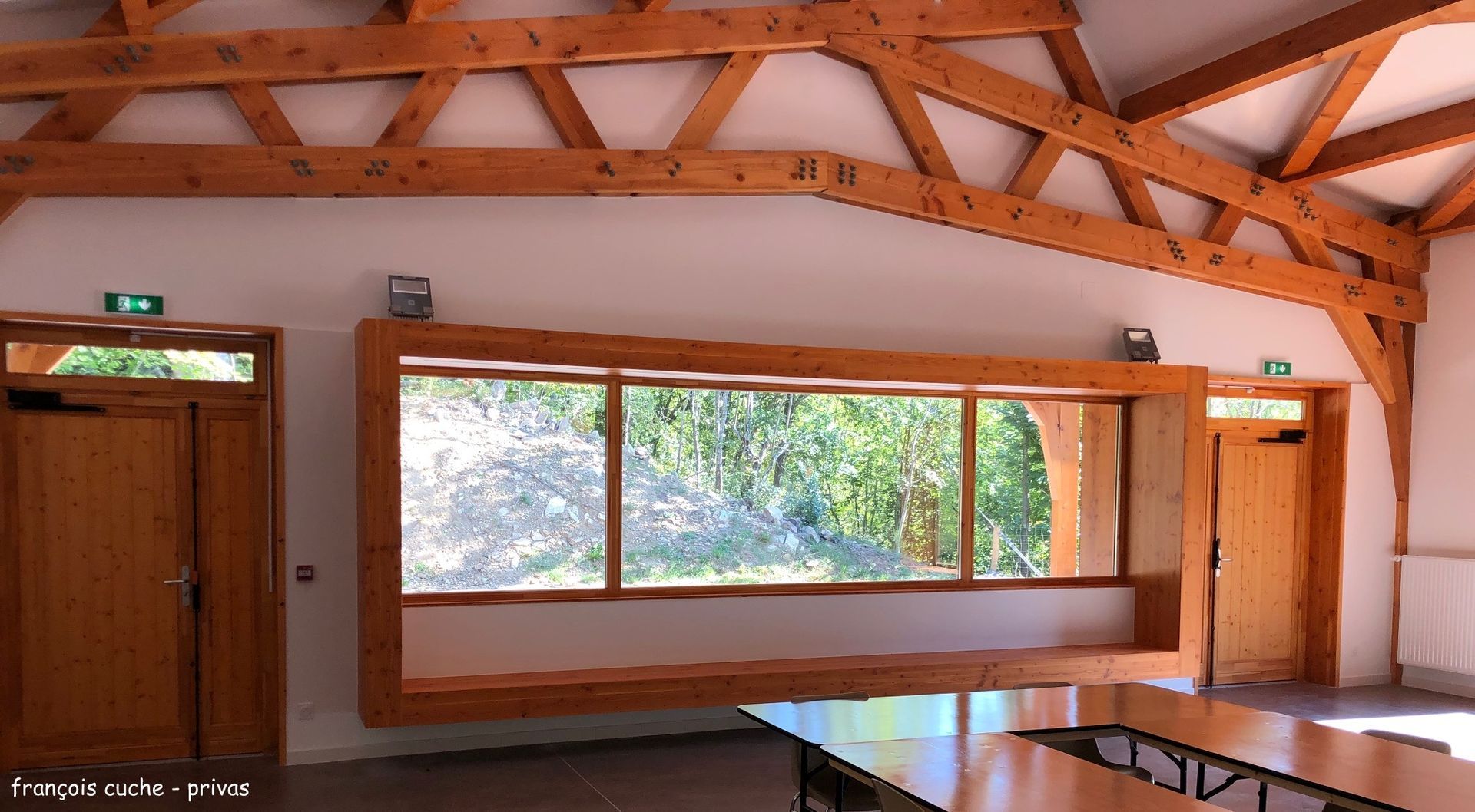Vue intérieure d'un bâtiment à poutres en bois avec une grande fenêtre et deux portes, dévoilant une vue sur la végétation verdoyante à l'extérieur.