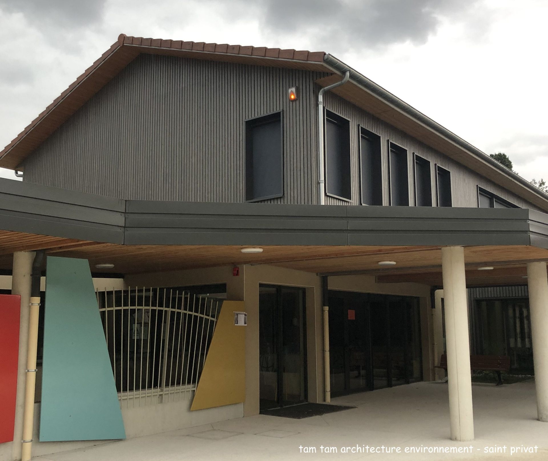 Bâtiment moderne avec bardage imitation bois, toit en surplomb, touches de couleur et colonnes.