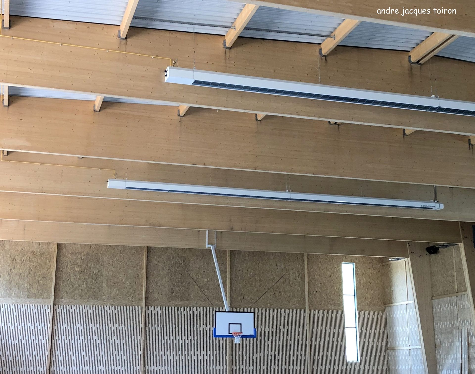 Intérieur d'un terrain de basket avec poutres en bois, panneau et éclairage blanc.