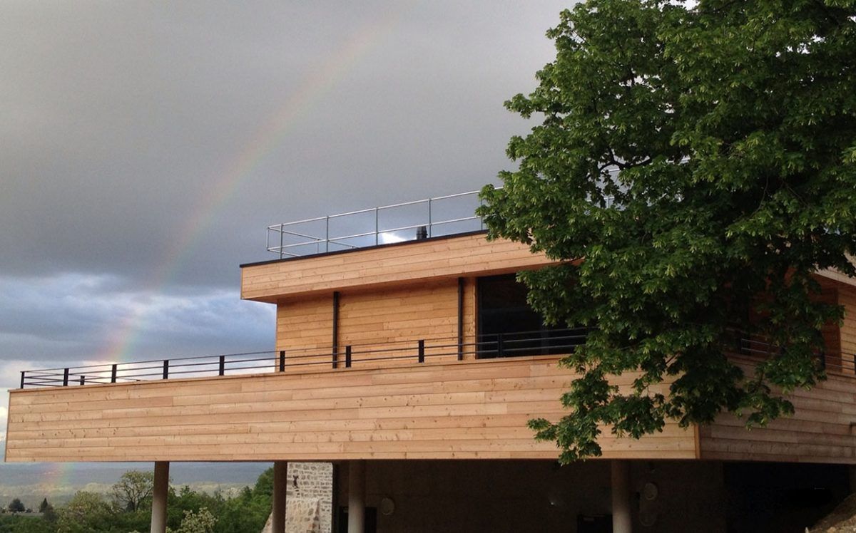 Maison en bois avec un arc-en-ciel partiel au-dessus et un arbre au premier plan.