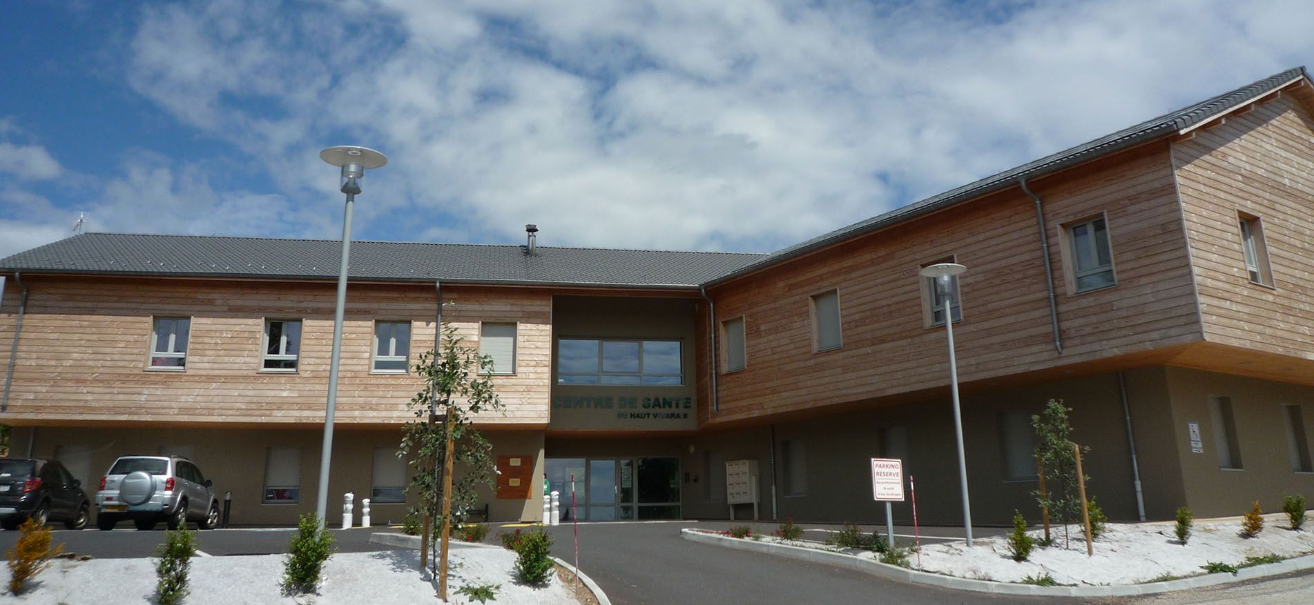 Extérieur d'un bâtiment moderne avec des panneaux de bois. Ciel bleu avec des nuages. Voitures garées au premier plan.