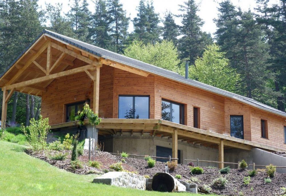 Maison en bois avec porche couvert, construite à flanc de colline et entourée d'arbres.
